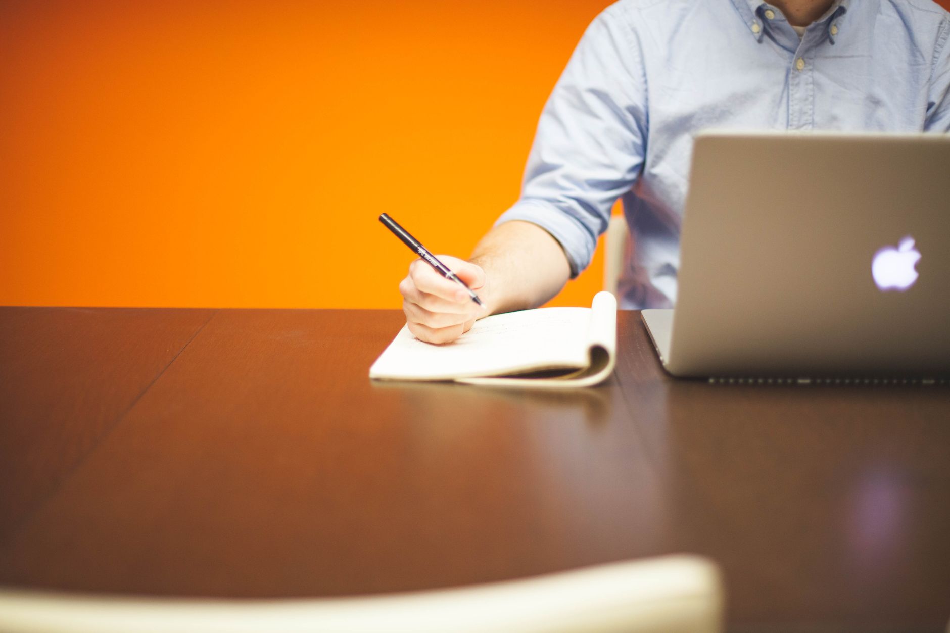 Adulta che scrive appunti su un quaderno accanto a un laptop, scrivania in legno con luce calda arancione.
