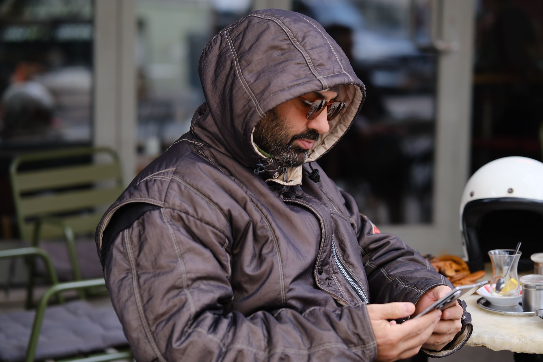 Uomo con cappuccio e occhiali da sole seduto al tavolino di un caffè all'aperto, mentre usa lo smartphone