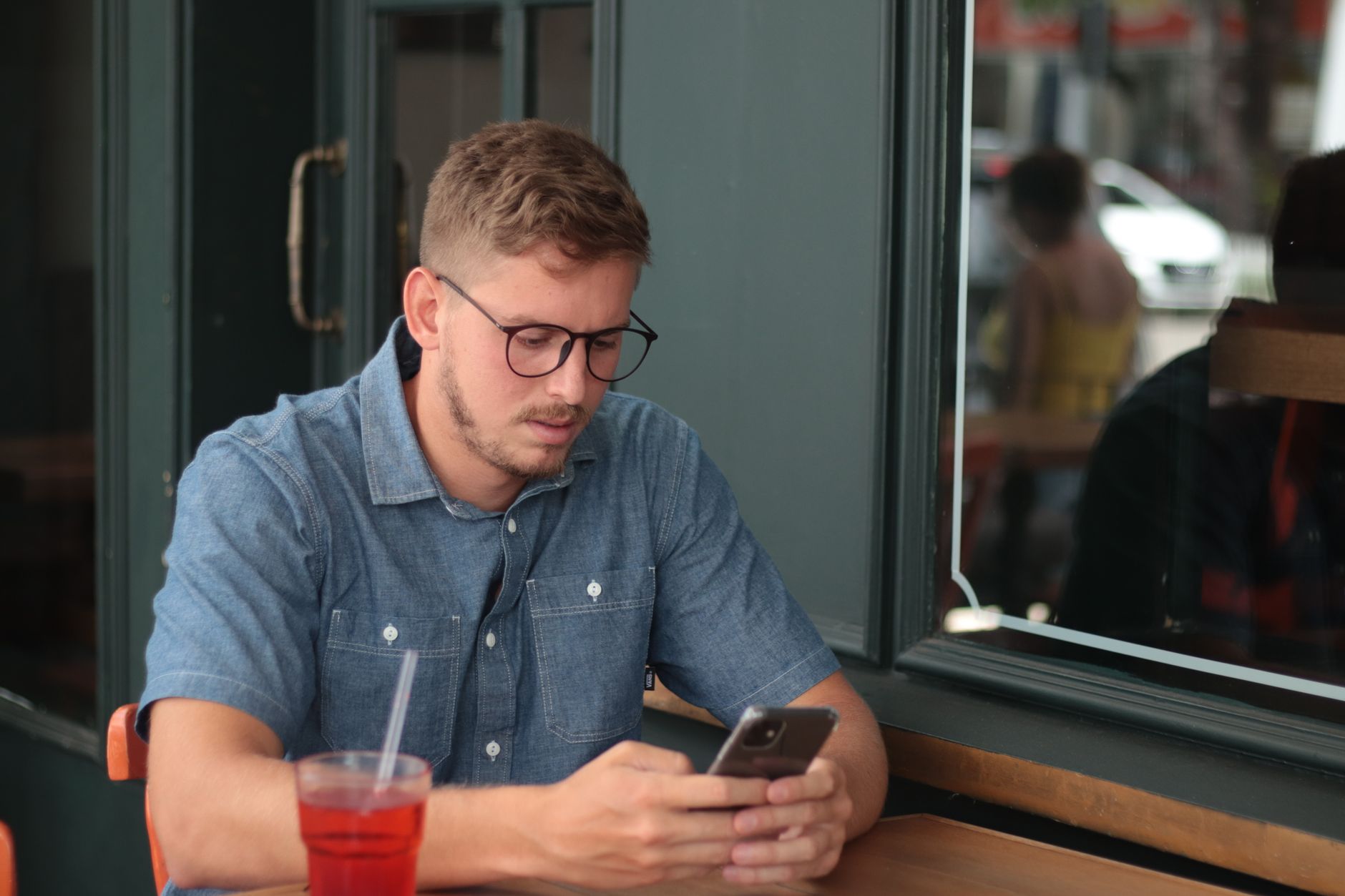 Giovane con occhiali e camicia di jeans seduto al tavolino di un bar, concentrato sullo smartphone, con una bibita rossa accanto